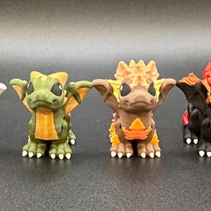 May include: A line of seven miniature dragon figures in various colours. The dragons are painted in shades of purple, white, green, brown, red, and yellow. Each has wings, small horns, and detailed features.