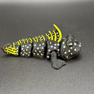May include: A black articulated figurine of a fish-like creature with a textured surface. The body features small white dots and bright yellow accents on the dorsal fin and tail. The figure has a segmented design allowing for movement.
