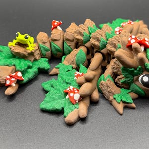 May include: A brown articulated creature figure with green leaf textures, small red and white spotted mushrooms, and a bright lime green frog perched on a rock. The figure has a textured bark-like appearance with large dark eyes and a segmented body.