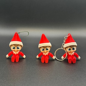 May include: Three small elf figurines. Each elf wears a red suit and hat with a white trim. The elves have blue eyes and are holding their arms out. One elf has a green wire attached to its hat, and another has a silver keychain.
