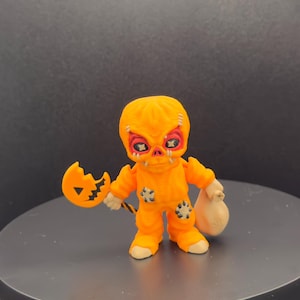 May include: A small, orange Halloween figurine of a character with a pumpkin-shaped head and stitched patches. The figure holds a broken pumpkin lollipop and a small beige sack. The figure has red eyes and a stitched mouth.