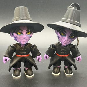 May include: Two 3D-printed figurines of a character with purple skin, yellow eyes, and a black outfit. Each figure wears a black hat and has a red belt. One figure has a keychain attachment. The figures are standing on a dark surface.