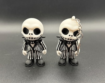 May include: Two Jack Skellington figurines. Each figure has a white head with large black eyes and a stitched mouth. They wear black and white striped suits and black shoes. One figure is a keychain.