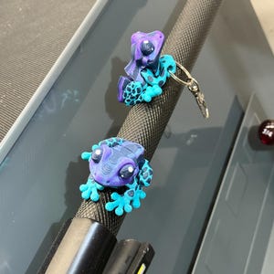 May include: Two gecko-shaped keychains. The geckos are primarily blue, purple, and black, with large, round eyes. One keychain is attached to a metal bar. The geckos have a textured appearance and are likely made of resin or a similar material.