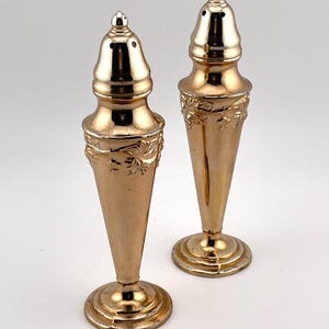 May include: A pair of tall, metallic gold-toned salt and pepper shakers with a conical body shape. Each shaker features a rounded, removable cap with small dispensing holes and an embossed floral vine pattern around the upper section.