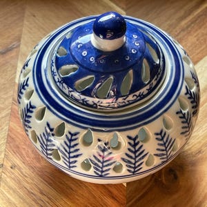 May include: A round white ceramic jar with a removable lid, featuring blue hand-painted fern leaf patterns and teardrop-shaped cutouts. The lid has a small blue knob handle and matching blue cutouts, creating a decorative openwork design.
