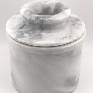 May include: A cylindrical butter keeper crafted from white and grey marbled stone. The container features a smooth, polished finish with a distinct lid that includes a central knob for easy handling, showcasing natural veining throughout the surface.