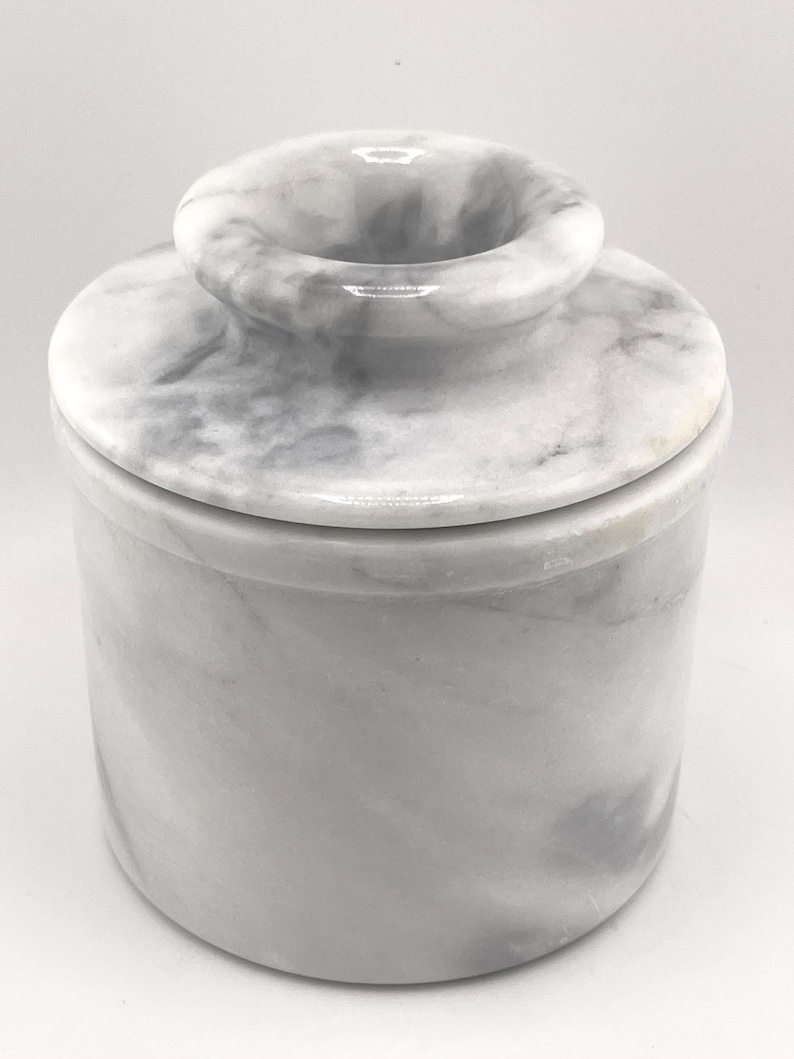 May include: A cylindrical butter keeper crafted from white and grey marbled stone. The container features a smooth, polished finish with a distinct lid that includes a central knob for easy handling, showcasing natural veining throughout the surface.
