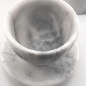 May include: A white marble cup with gray veining resting on a matching circular marble saucer. The cup has a smooth, rounded interior and a thick rim, while the saucer features a flat surface with a subtle mottled pattern throughout the stone.
