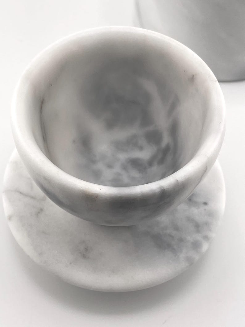 May include: A white marble cup with gray veining resting on a matching circular marble saucer. The cup has a smooth, rounded interior and a thick rim, while the saucer features a flat surface with a subtle mottled pattern throughout the stone.