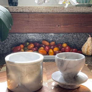 May include: A cylindrical white marble container with grey veining sits next to a smaller, rounded white marble cup with a wide base and grey veining. Both items are crafted from polished stone and feature natural, unique grey marble patterns.