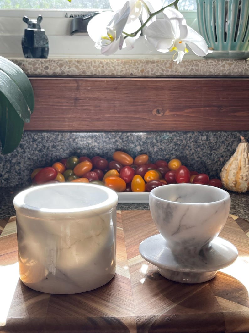 May include: A cylindrical white marble container with grey veining sits next to a smaller, rounded white marble cup with a wide base and grey veining. Both items are crafted from polished stone and feature natural, unique grey marble patterns.