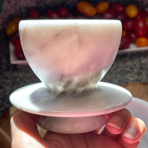 May include: A white marble cup with subtle grey veining, featuring a wide, rounded bowl shape that tapers down to a narrow stem and a circular base. The stone surface has a smooth, polished finish with natural variations in color and texture.