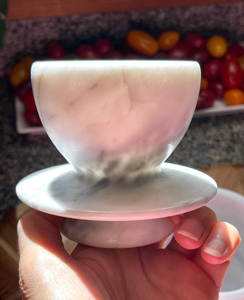 May include: A white marble cup with subtle grey veining, featuring a wide, rounded bowl shape that tapers down to a narrow stem and a circular base. The stone surface has a smooth, polished finish with natural variations in color and texture.