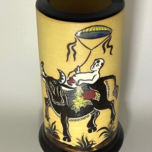 May include: A cylindrical ceramic vase with a yellow base and black rim and base. The exterior features a hand-painted illustration of a person riding a black ox, holding a string attached to a floating blue and yellow umbrella-like object.