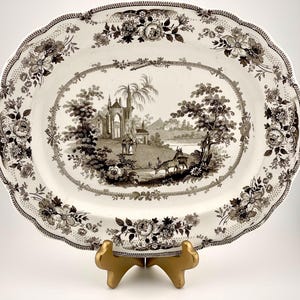 May include: An oval ceramic platter featuring a dark brown transferware pattern on a cream background. The center depicts a landscape with a gothic structure, trees, and deer. The scalloped rim is decorated with a detailed floral border.