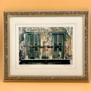 Könnte beinhalten: Ein gerahmtes Foto mit dem Titel Verona Balcony von Tony Casper, das zwei Fenster mit grünen Fensterläden und einen schmiedeeisernen Balkon mit Topfblumen vor einer verwitterten, strukturierten Wand in einem goldenen Rahmen zeigt.