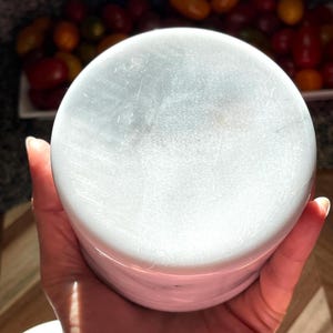 May include: A cylindrical white marble container with subtle grey veining throughout its surface. The object features a smooth, polished finish and a flat circular top, showcasing the natural stone texture and variations in the white and grey color palette.