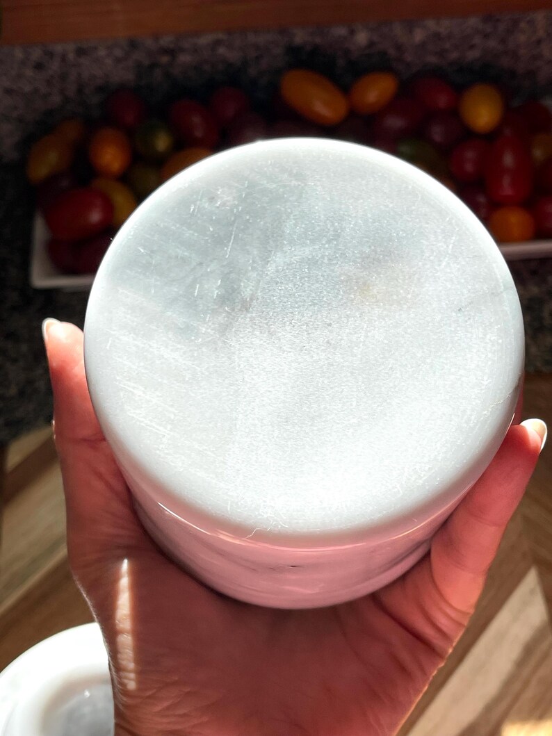 May include: A cylindrical white marble container with subtle grey veining throughout its surface. The object features a smooth, polished finish and a flat circular top, showcasing the natural stone texture and variations in the white and grey color palette.