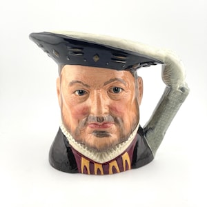 May include: A ceramic character jug shaped like the head and shoulders of a bearded man wearing a black Tudor-style hat with a white feather. The figure wears a black doublet with gold accents over a white ruff collar, with a gray stone-textured handle.