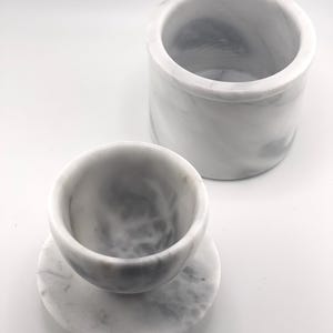 May include: A white marble container with grey veining and a matching white marble cup resting on a circular marble base. The cylindrical container is open at the top, and the cup features a smooth, rounded interior with natural stone patterns.