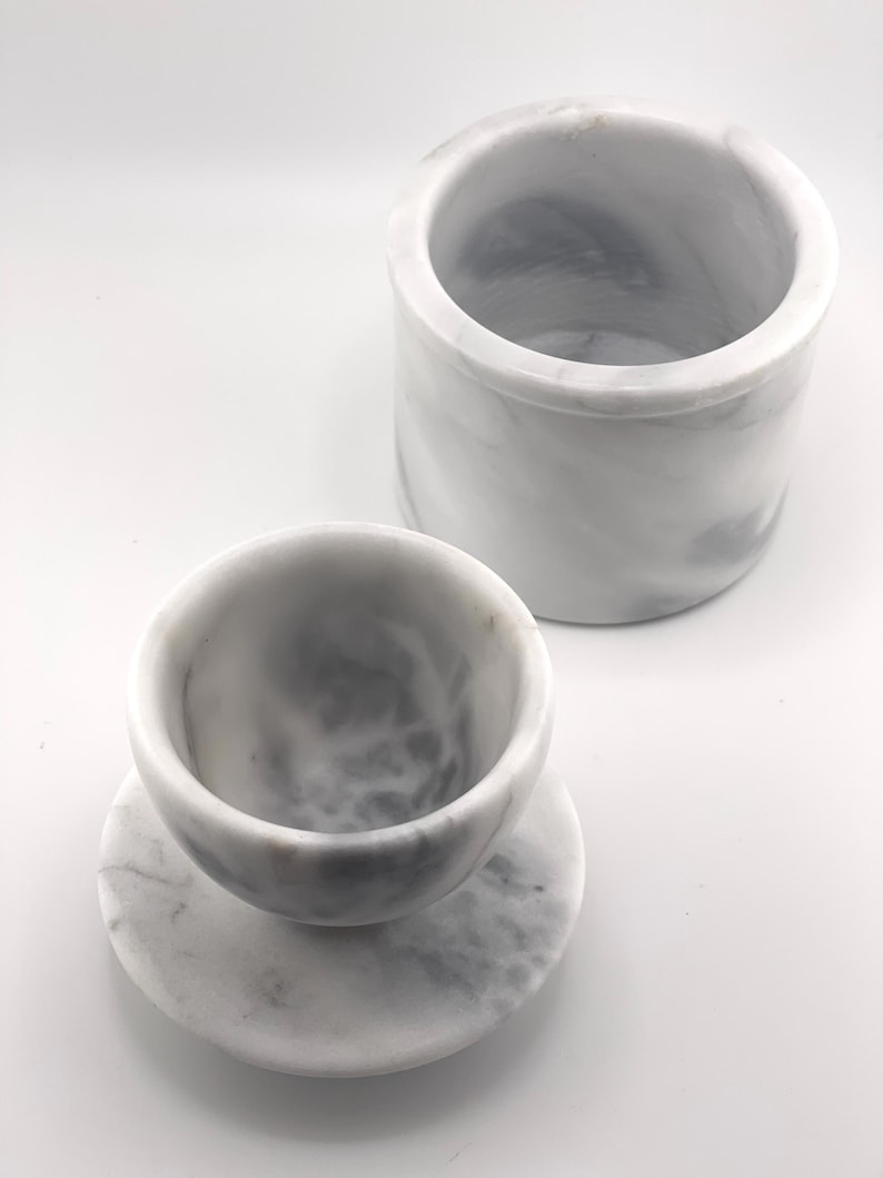 May include: A white marble container with grey veining and a matching white marble cup resting on a circular marble base. The cylindrical container is open at the top, and the cup features a smooth, rounded interior with natural stone patterns.