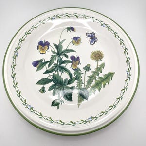 May include: A round ceramic plate with a cream-colored base and a green rim. The center features a botanical illustration of purple and yellow pansies, a yellow dandelion, and a small blue butterfly. A delicate green vine border circles the edge.