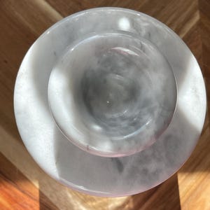 May include: A circular white marble mortar and pestle set viewed from directly above. The mortar features a smooth, polished surface with natural grey veining throughout the stone. The pestle is centered within the bowl, showcasing a matching white and grey marbled pattern with a rounded, polished head.