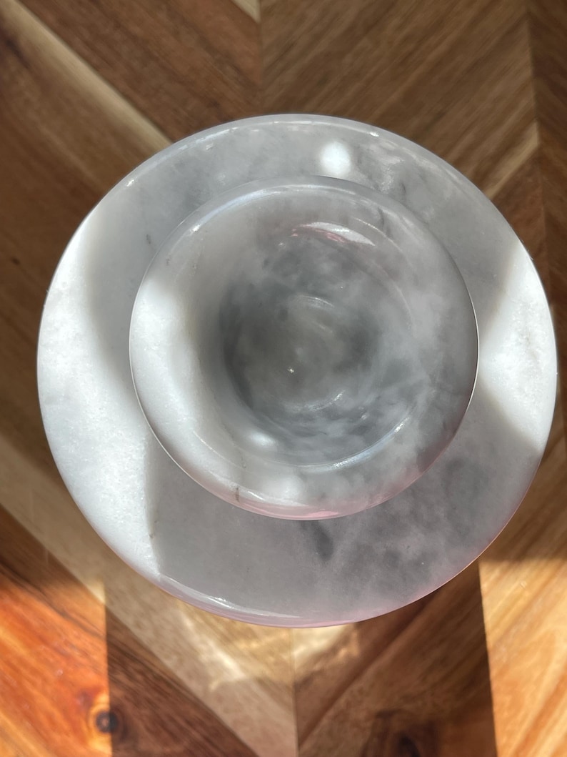 May include: A circular white marble mortar and pestle set viewed from directly above. The mortar features a smooth, polished surface with natural grey veining throughout the stone. The pestle is centered within the bowl, showcasing a matching white and grey marbled pattern with a rounded, polished head.