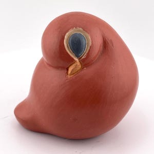 May include: A smooth, terracotta-colored ceramic bird figurine with a rounded, compact body. The bird features a single visible eye painted in dark blue with a light tan outline, and a small, pointed, light tan beak tucked against its side.