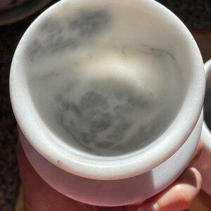 May include: A white marble cup with a smooth, rounded rim and a deep, cylindrical interior. The stone features natural grey veining patterns that swirl across the inner surface, creating a unique, mottled appearance within the polished bowl.