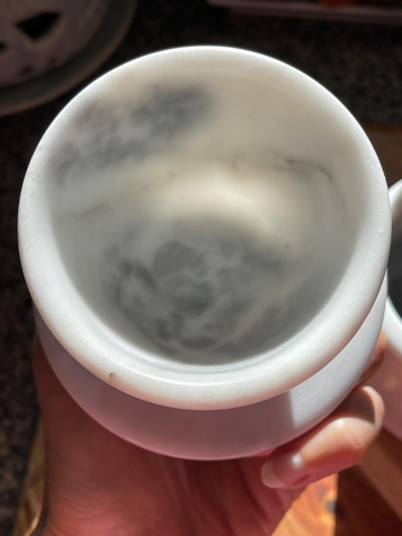 May include: A white marble cup with a smooth, rounded rim and a deep, cylindrical interior. The stone features natural grey veining patterns that swirl across the inner surface, creating a unique, mottled appearance within the polished bowl.