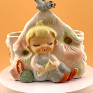 May include: A ceramic planter featuring a sculpted figure of a blonde child in blue overalls sitting against a white structure. A small blue bird is perched on top. The child has closed eyes and holds a yellow object. Accents include green and orange.