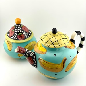 May include: A ceramic teapot and a matching lidded sugar bowl featuring a bright turquoise base color. The teapot has a yellow grid-patterned lid, a black and white striped handle, and yellow banana illustrations. The sugar bowl has a strawberry lid.