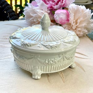 May include: A round, off-white ceramic lidded dish featuring a raised floral garland pattern, vertical fluting along the base, and three small feet. The lid is topped with a pointed, textured finial and displays a radiating sunburst design.