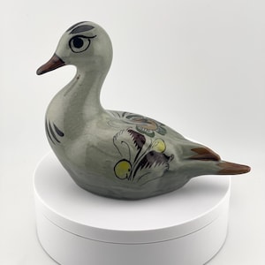 May include: A ceramic duck figurine with a glossy light gray finish. The duck features hand-painted floral motifs in shades of yellow, brown, and green on its back and sides, with dark brown accents on the beak, eyes, and tail feathers.