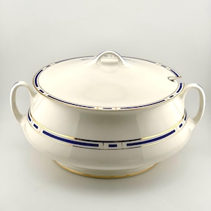 May include: A cream-colored ceramic soup tureen with a matching lid, featuring two side handles and a small notch for a ladle. The exterior is decorated with a horizontal band of deep blue and gold trim, which is also repeated on the lid.