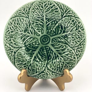 May include: A round ceramic plate featuring a raised, embossed green cabbage leaf pattern with intricate veining radiating from the center. The plate is displayed upright on a small, gold-colored metal stand with three curved support legs.