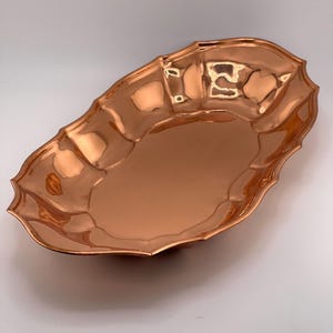 Op de afbeelding: Een ovale koperen serveerschaal met een gepolijst, reflecterend oppervlak. De schaal heeft een geschulpt, golvend randontwerp dat een reeks afgeronde inkepingen langs de omtrek creëert, wat zorgt voor een opvallend en getextureerd uiterlijk.