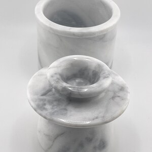 May include: A white marble butter keeper with gray veining, consisting of a cylindrical base container and a separate lid featuring a central handle. The stone surface displays natural patterns and a smooth, polished finish throughout the piece.
