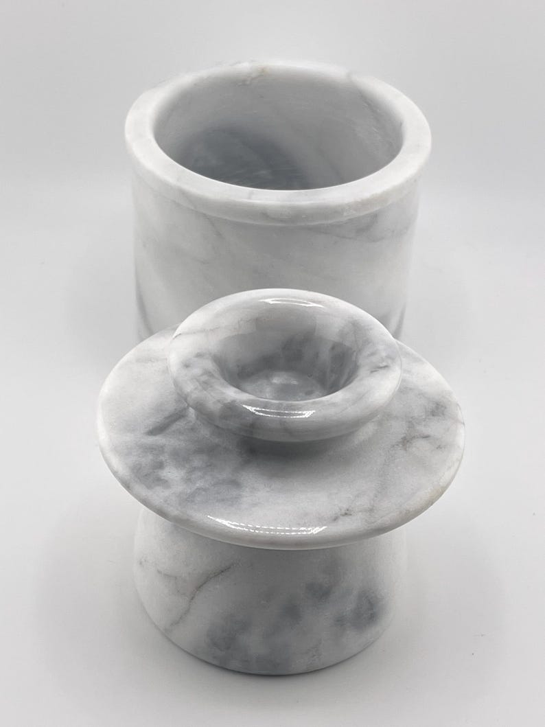 May include: A white marble butter keeper with gray veining, consisting of a cylindrical base container and a separate lid featuring a central handle. The stone surface displays natural patterns and a smooth, polished finish throughout the piece.