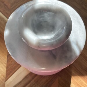 May include: A circular white and gray marble container with a matching lid. The lid features a raised, rounded handle in the center. The stone surface displays natural veining patterns in shades of gray against a white background, creating a smooth, polished appearance from a top-down perspective.