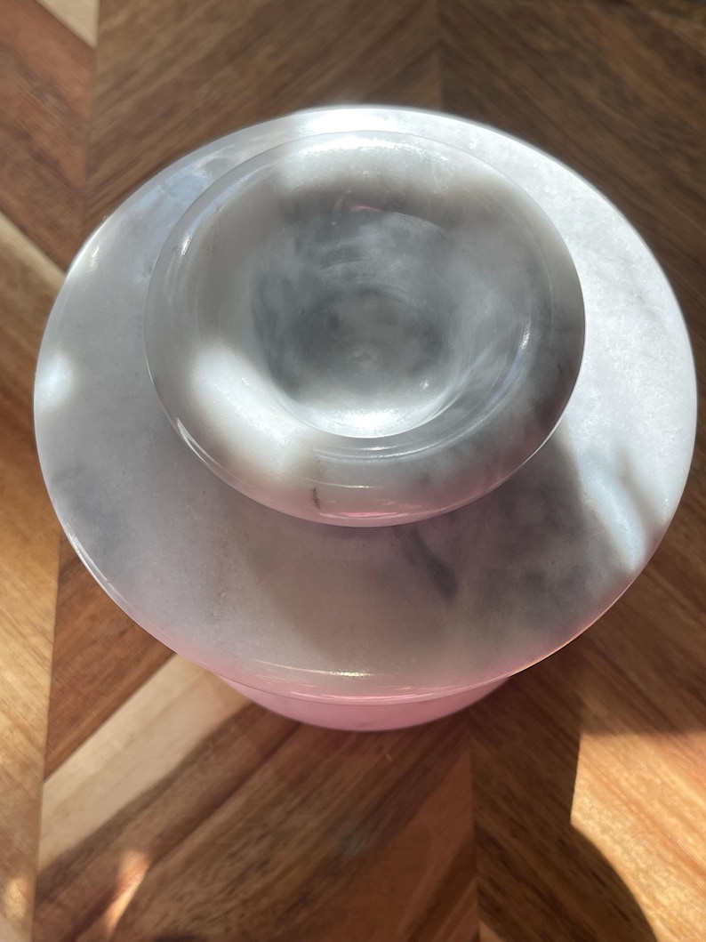 May include: A circular white and gray marble container with a matching lid. The lid features a raised, rounded handle in the center. The stone surface displays natural veining patterns in shades of gray against a white background, creating a smooth, polished appearance from a top-down perspective.