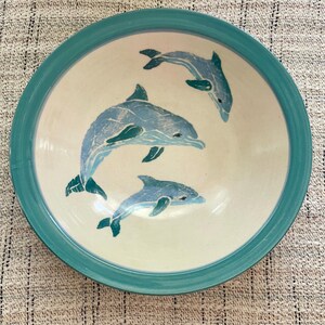 May include: A round ceramic bowl with a cream-colored interior and a wide teal rim. The interior features three blue dolphin illustrations of varying sizes, depicted in a stylized, textured print style, swimming in a circular arrangement.