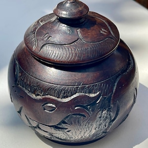 May include: A dark brown, spherical wooden jar with a matching lid. The exterior surface features intricate, hand-carved relief patterns depicting stylized animal figures and textured foliage, with a small, rounded knob handle on the lid.