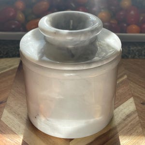 May include: A cylindrical white marble butter keeper with a smooth, polished finish and subtle grey veining. The container consists of a base and a removable lid with a central knob, featuring a classic design crafted from natural stone material.