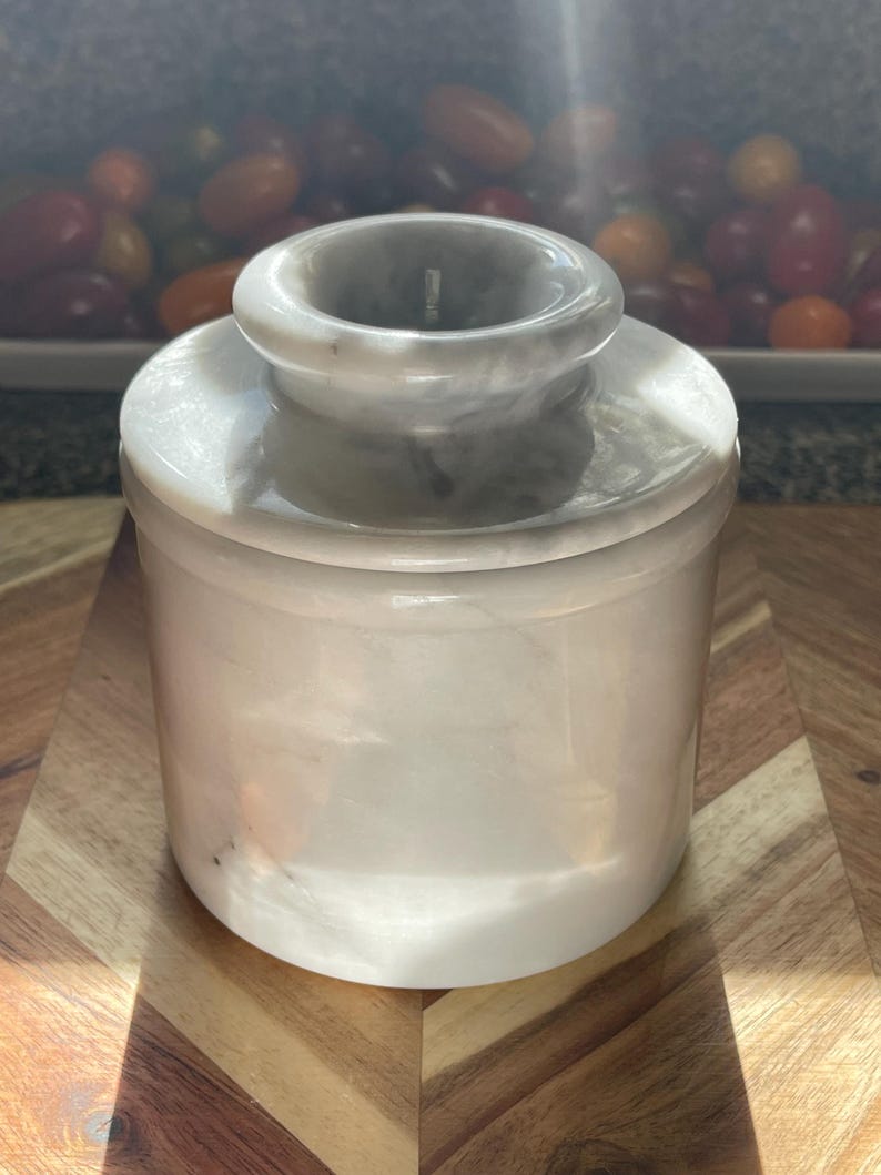 May include: A cylindrical white marble butter keeper with a smooth, polished finish and subtle grey veining. The container consists of a base and a removable lid with a central knob, featuring a classic design crafted from natural stone material.