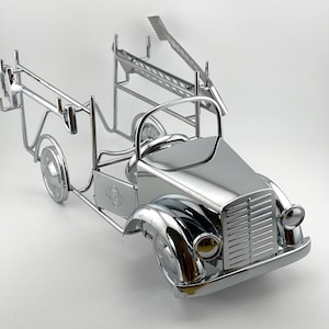 May include: A shiny chrome-finished metal sculpture shaped like a vintage fire truck. The model features a detailed front grille, round headlights, a steering wheel, and an open-frame rear section with a ladder attachment extending from the back.