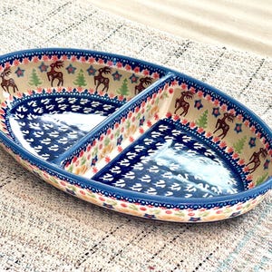 May include: An oval ceramic serving dish with a central divider, featuring a hand-painted pattern of brown reindeer and green pine trees on a cream background, accented by a deep blue border with white floral motifs and red and blue trim.