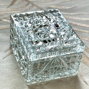 May include: A square clear glass trinket box featuring a multifaceted geometric pattern across the lid and sides. The glass surface is deeply cut with intersecting lines and diamond shapes, creating a reflective and textured appearance throughout.